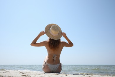 Woman in bikini sitting on sandy beach near sea, back view