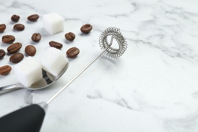 Black milk frother wand, sugar cubes and coffee beans on white marble table, closeup. Space for text