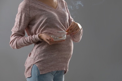 Photo of Young pregnant woman smoking cigarette on grey background, closeup. Space for text