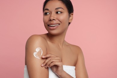 Young woman applying body cream onto arm on pink background