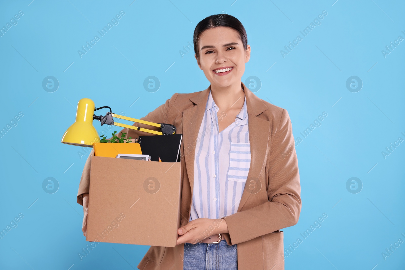 Photo of Happy unemployed woman with box of personal office belongings on light blue background