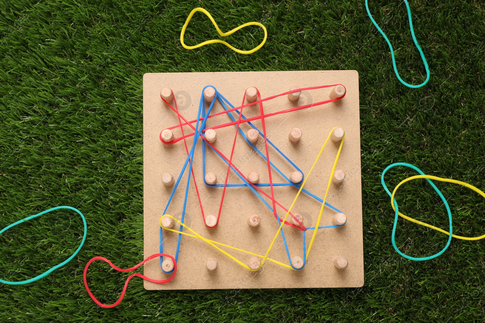 Photo of Wooden geoboard with rubber bands on artificial grass, flat lay. Educational toy for motor skills development