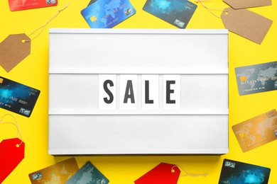 Photo of Lightbox with word Sale, credit cards and price tags on yellow background, flat lay