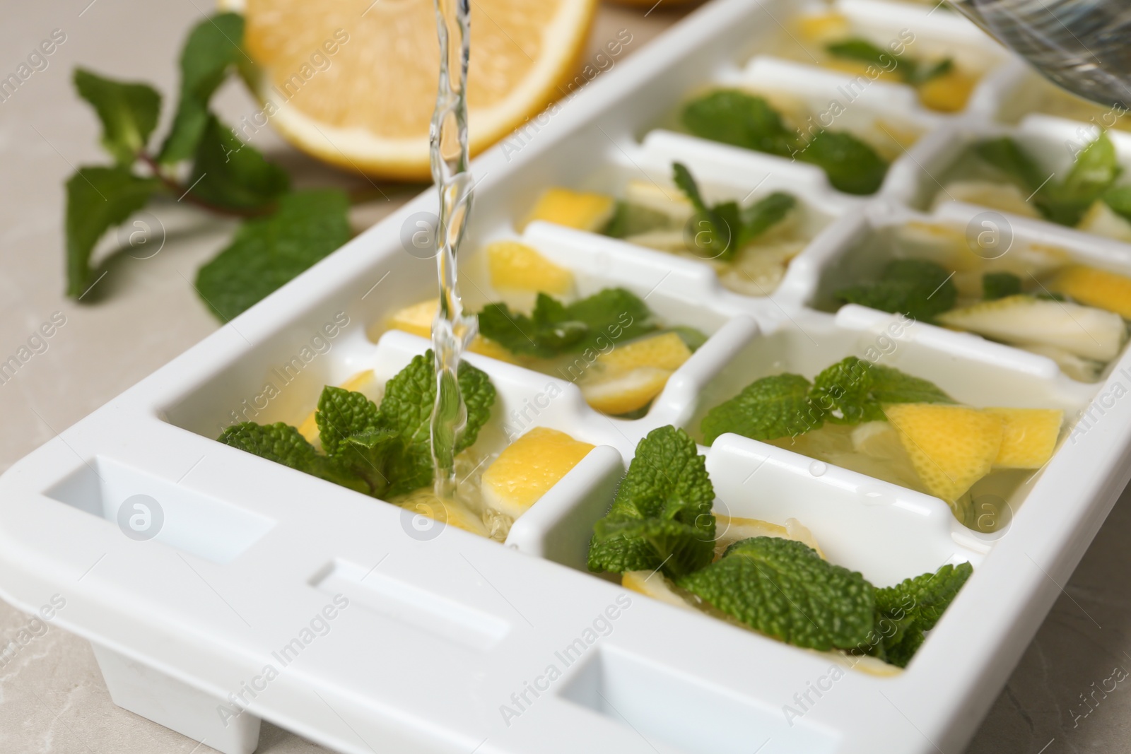 Photo of Pouring water into ice cube tray with mint and lemon on table, closeup