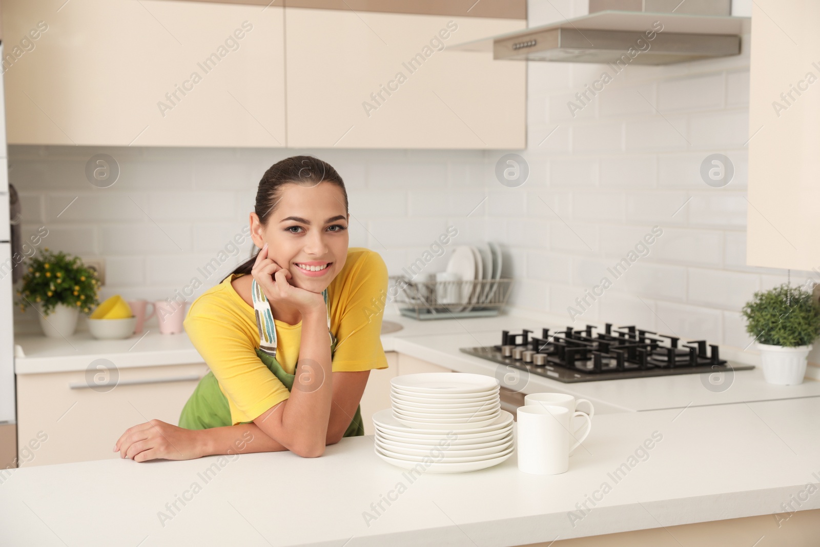 Photo of Beautiful young woman with clean dishes and cups at table in kitchen