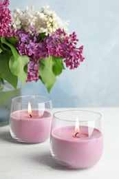 Burning wax candles in glass holders and lilac flowers on white table, space for text