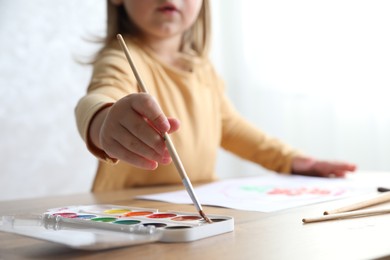 Photo of Little girl drawing with brush at wooden table indoors, selective focus. Child`s art