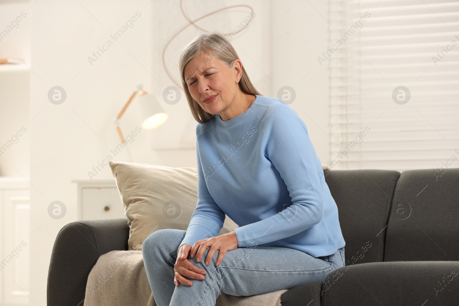 Photo of Mature woman suffering from knee pain on sofa at home. Rheumatism symptom