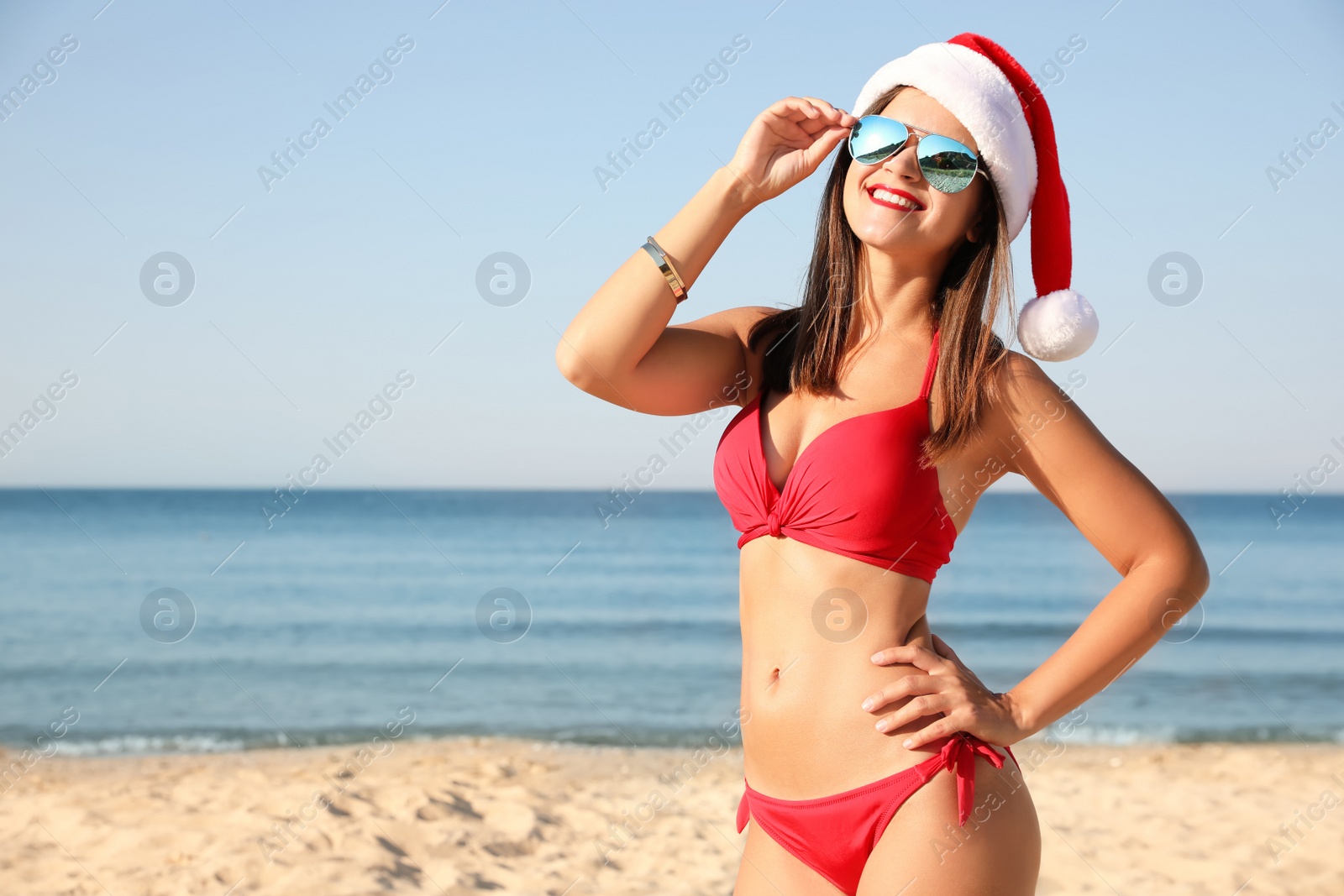 Photo of Young woman wearing Santa hat and bikini on beach, space for text. Christmas vacation