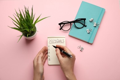 Photo of Woman writing in notebook on pink background, top view. New Year aims