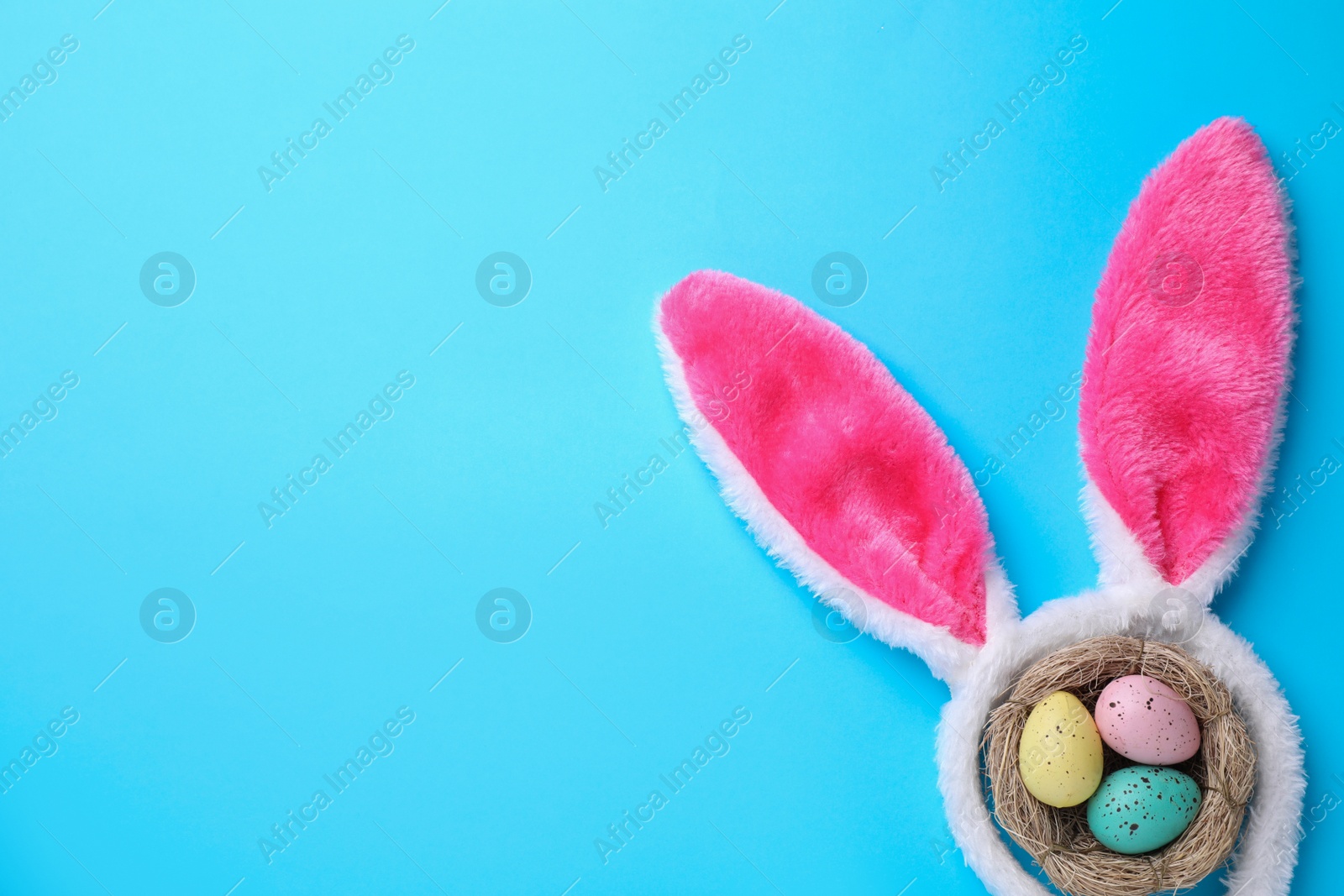 Photo of Headband with Easter bunny ears and dyed eggs in nest on light blue background, flat lay. Space for text