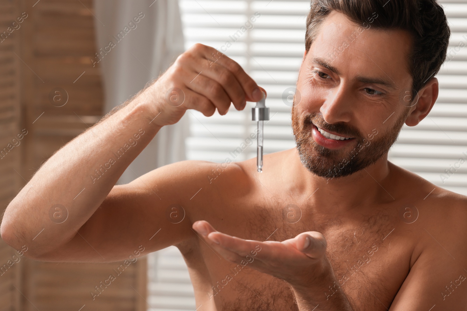 Photo of Smiling man dropping cosmetic serum onto his hand indoors