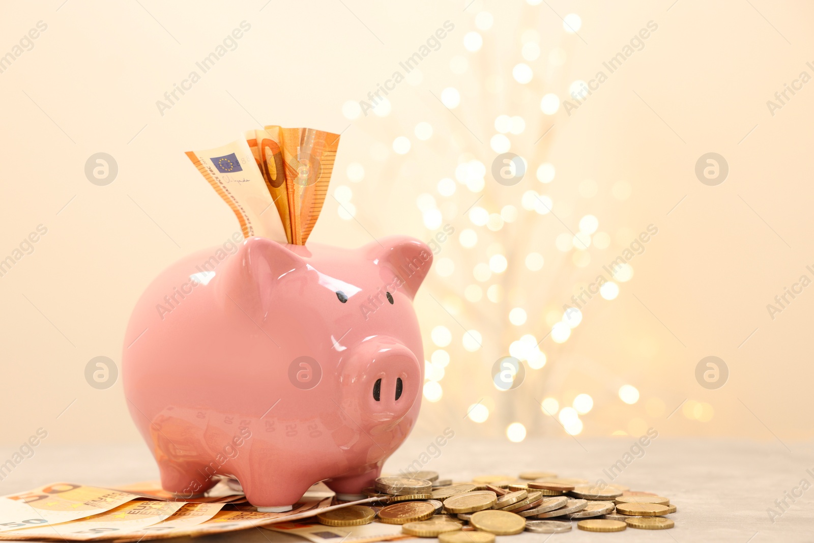 Photo of Piggy bank with euro banknotes and coins on grey table against blurred lights, space for text