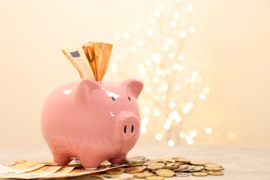 Photo of Piggy bank with euro banknotes and coins on grey table against blurred lights, space for text