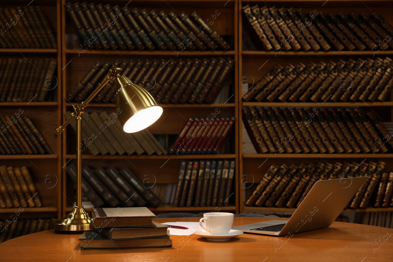 Photo of Lamp, cup of drink and laptop on wooden table near shelves with collection of vintage books in library