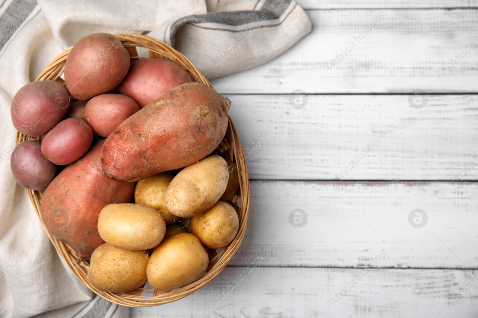 Photo of Different types of fresh potatoes in wicker basket on white wooden table, flat lay. Space for text