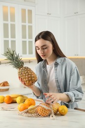 Photo of Woman with pineapple and string bag of fresh fruits at light marble table in kitchen