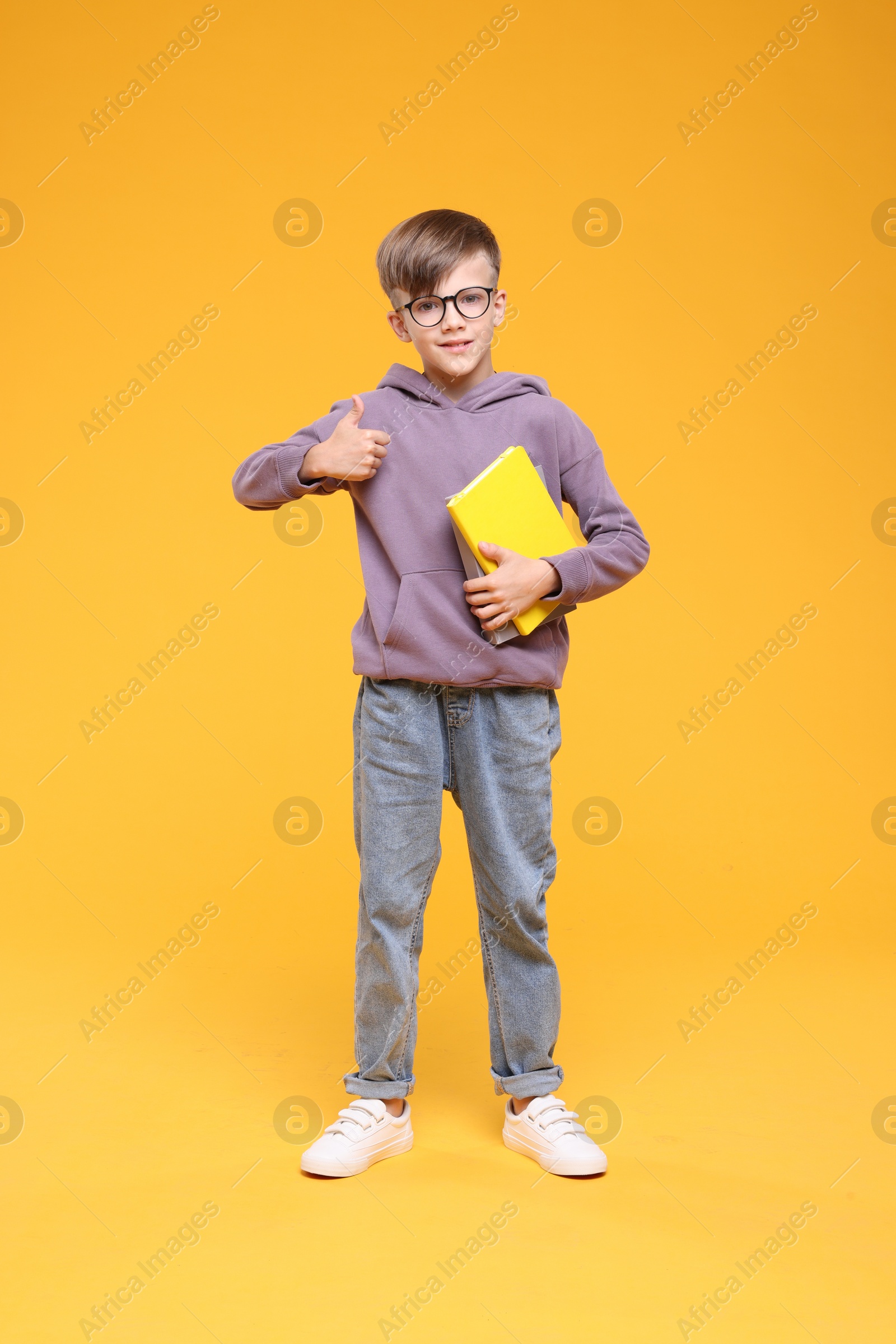 Photo of Cute schoolboy in glasses holding books and showing thumbs up on orange background