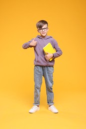 Cute schoolboy in glasses holding books and showing thumbs up on orange background