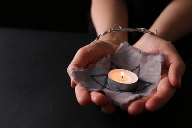 Photo of Woman tied with barbed wire holding star of David and burning candle on black background, closeup. Holocaust memory day