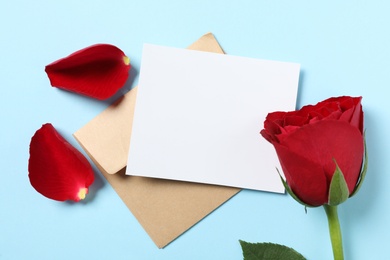 Photo of Blank greeting card, envelope and rose on light blue background, flat lay. Valentine's day celebration