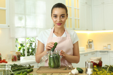 Woman putting cucumber into pickling jar at table in kitchen