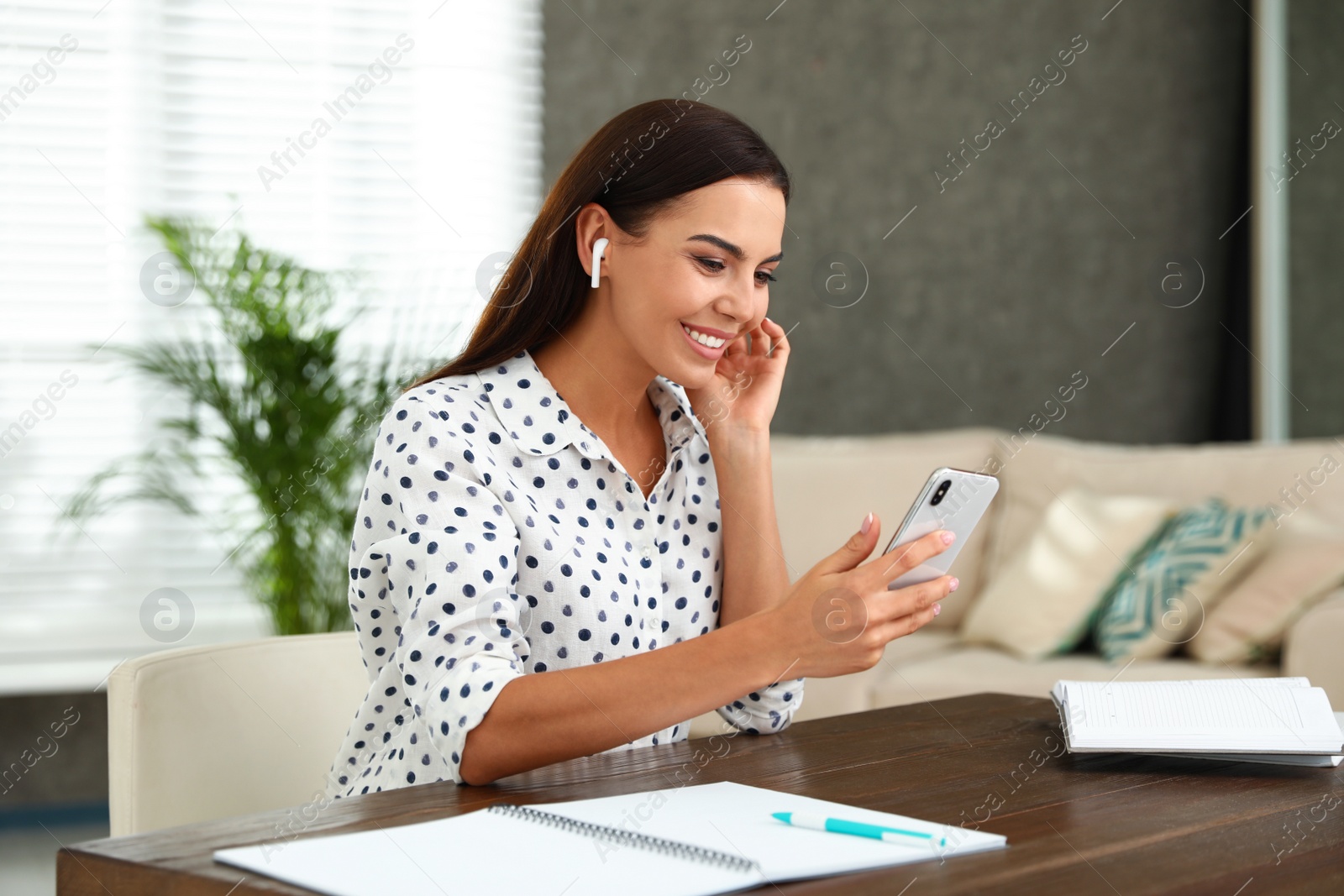 Photo of Happy young woman with smartphone listening to music through wireless earphones at table in office