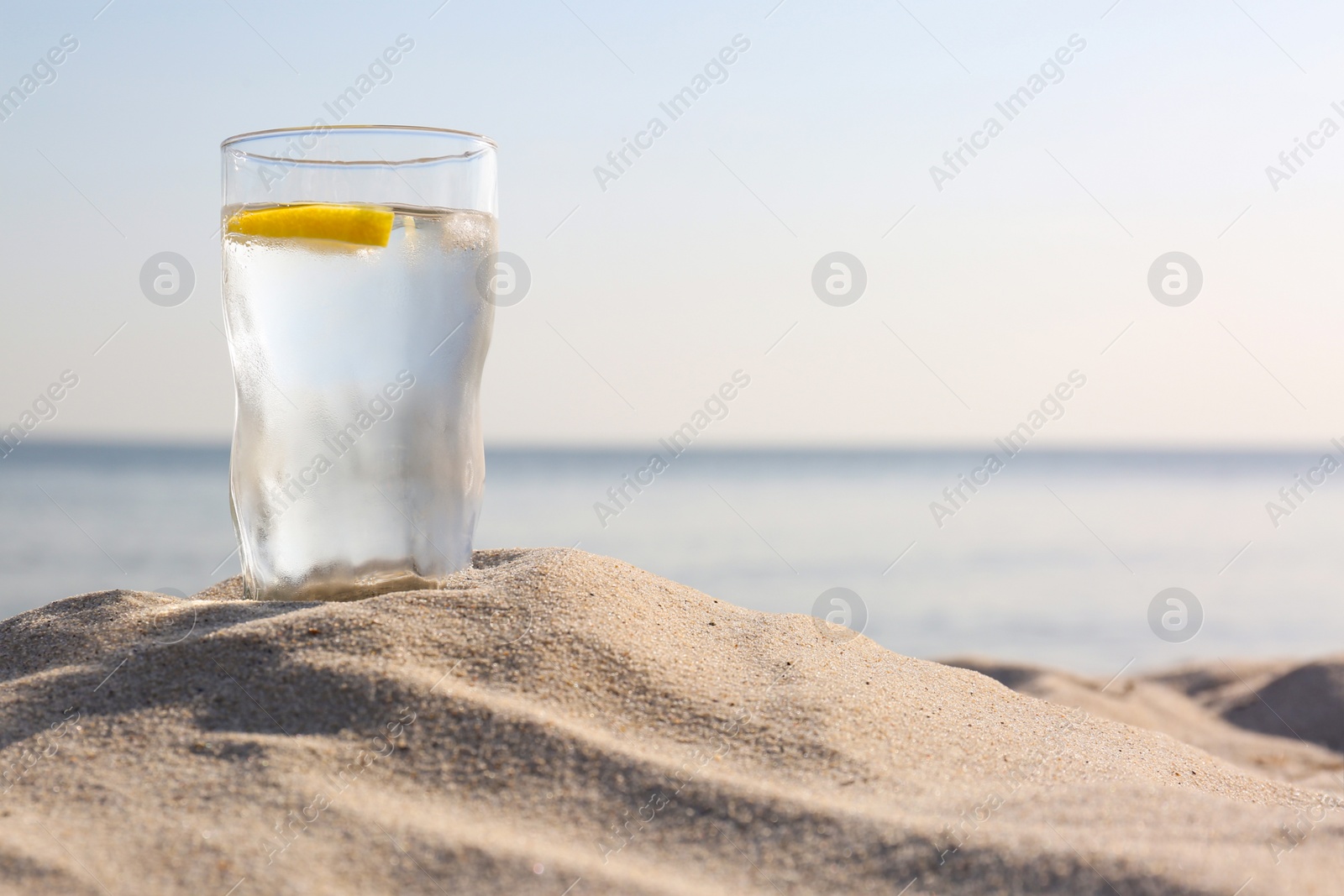 Photo of Sandy beach with glass of refreshing lemon drink on hot summer day, space for text