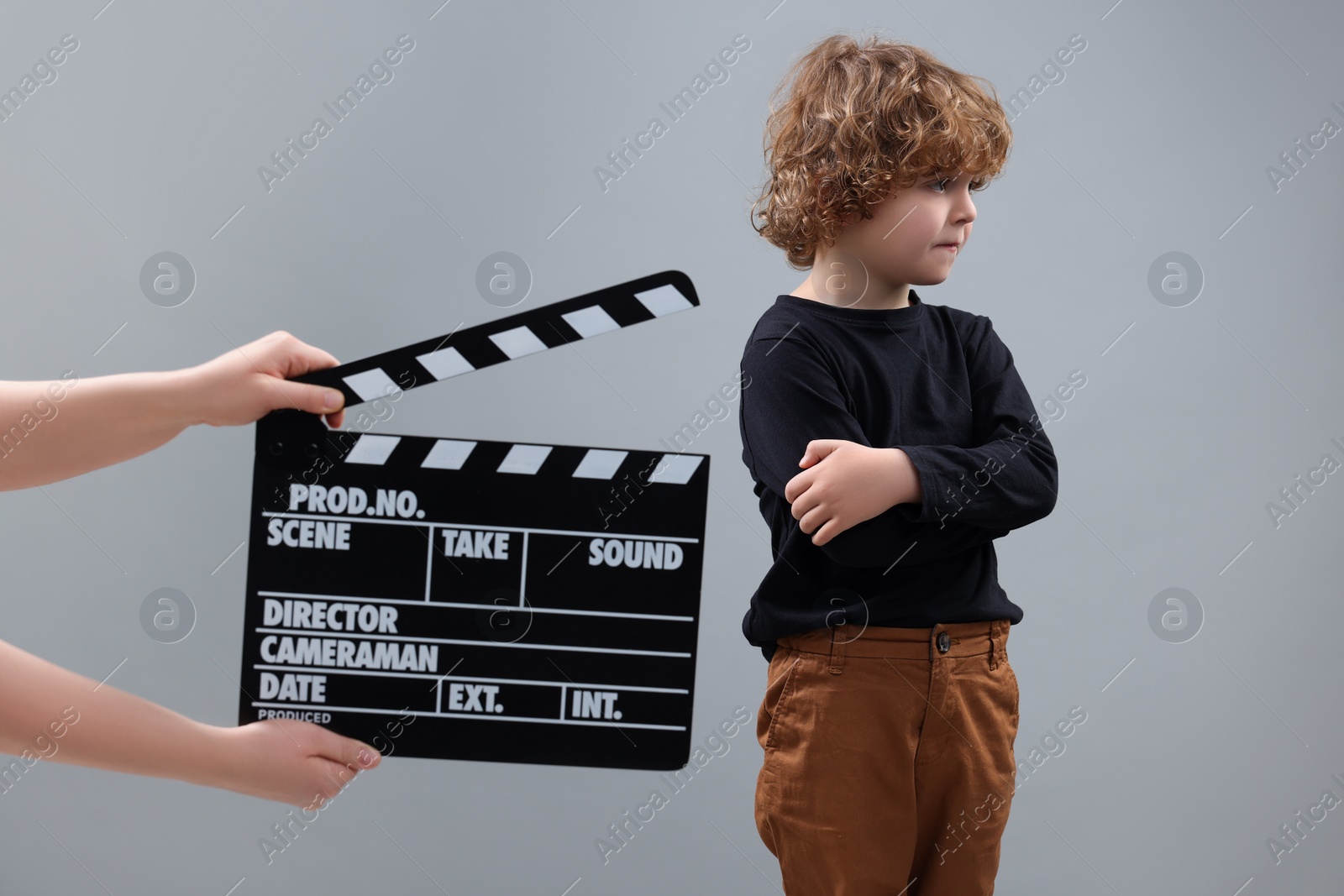 Photo of Sad boy performing while second assistant camera holding clapperboard on grey background. Little actor