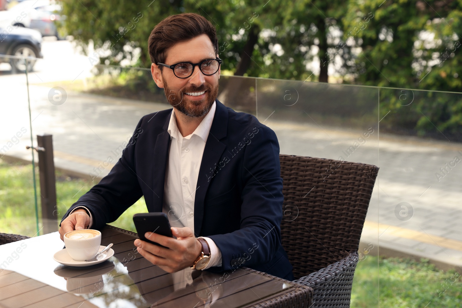 Photo of Handsome bearded man with cup of drink and smartphone at table in outdoor cafe