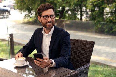 Photo of Handsome bearded man with cup of drink and smartphone at table in outdoor cafe