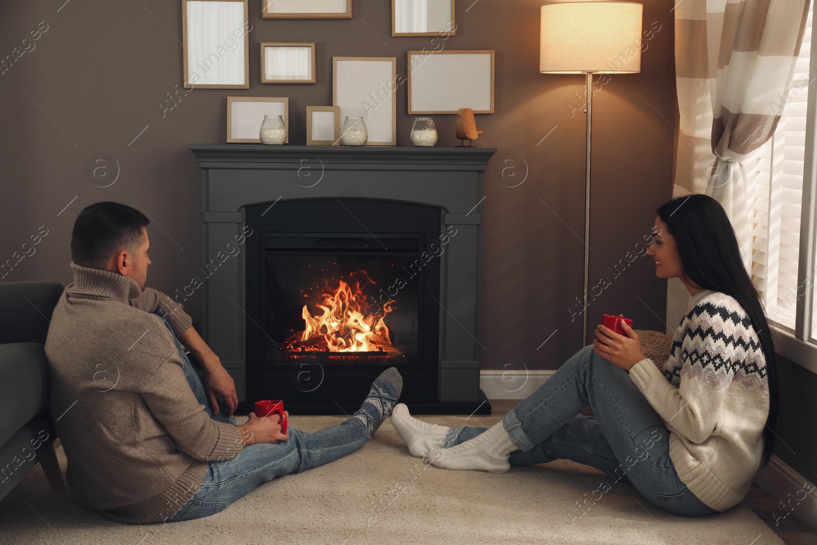 Photo of Lovely couple with cups of hot drink spending time together near fireplace at home