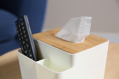 Box with paper tissues and TV remote control on wooden table in living room, closeup