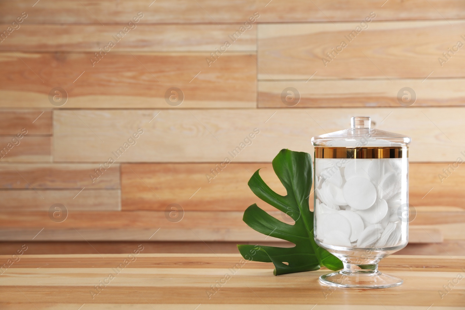 Photo of Decorative glass jar with cotton pads and monstera leaf on table against wooden background. Space for text