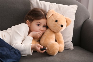 Little girl with toy bear changing TV channels by remote control on sofa at home