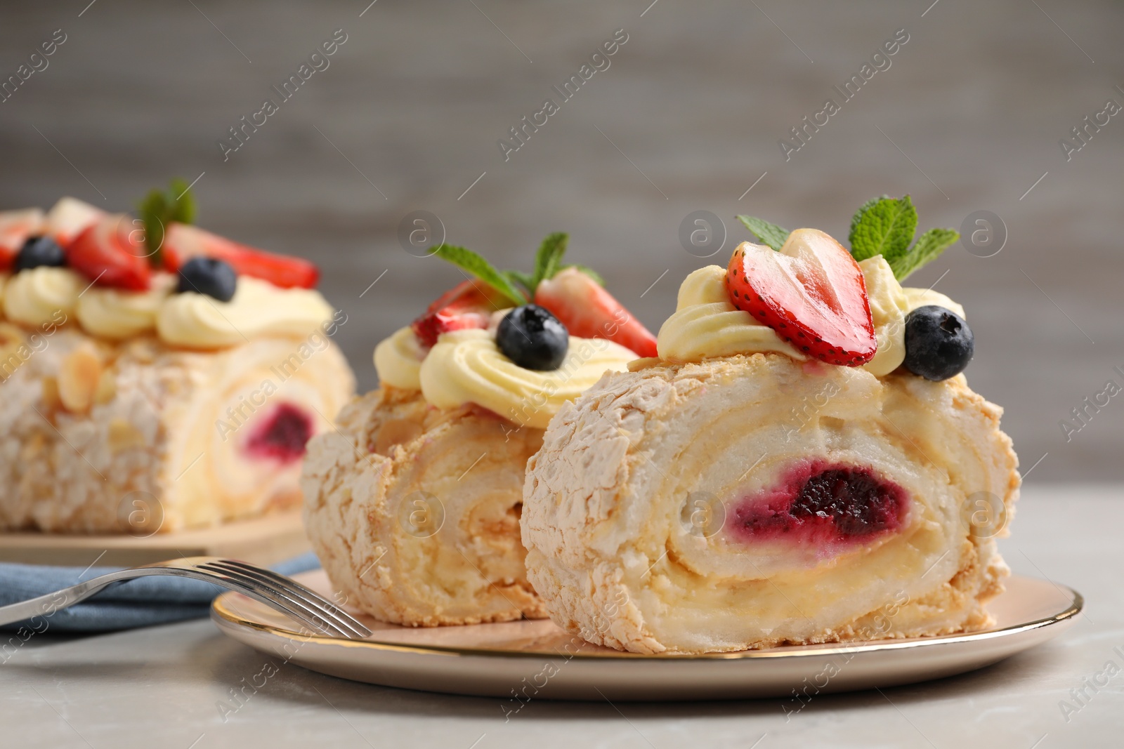 Photo of Pieces of tasty meringue roll with jam, cream, strawberry, blueberry and mint on light grey table, closeup