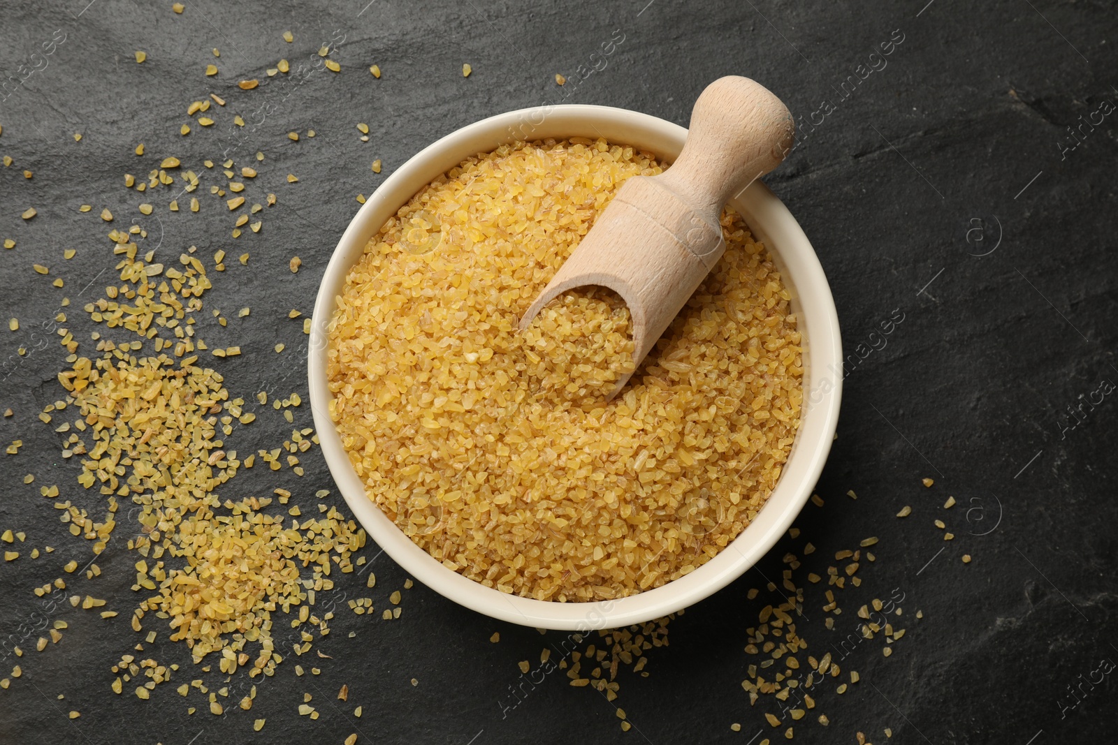 Photo of Bowl and scoop with raw bulgur on dark gray table, top view