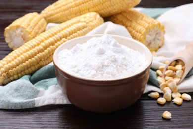 Photo of Bowl with corn starch, ripe cobs and kernels on dark wooden table, closeup