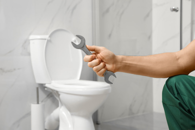 Photo of Professional plumber holding wrench near toilet bowl in bathroom, closeup