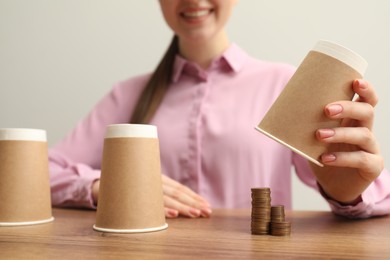 Shell game. Woman showing stack of coins under cup at wooden table, closeup