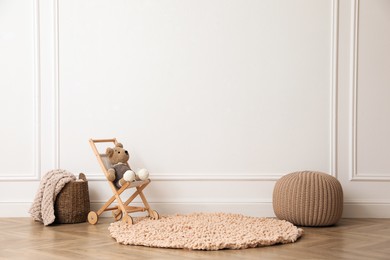 Photo of Toy stroller with bear, pouf and wicker basket near white wall in child room. Interior design