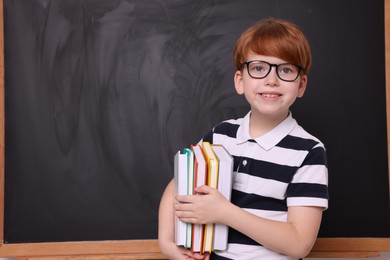 Photo of Smiling schoolboy in glasses with books near blackboard. Space for text