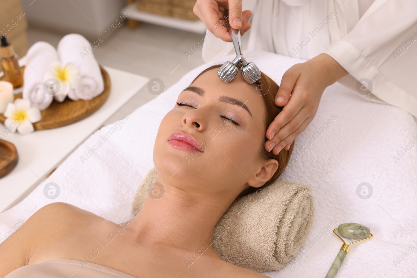 Photo of Young woman receiving facial massage with metal roller in beauty salon, closeup