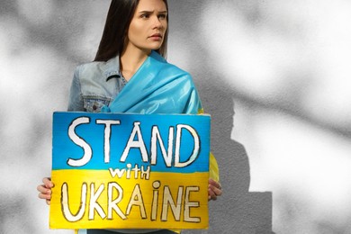 Photo of Sad woman holding poster in colors of national flag and words Stand with Ukraine near light wall. Space for text