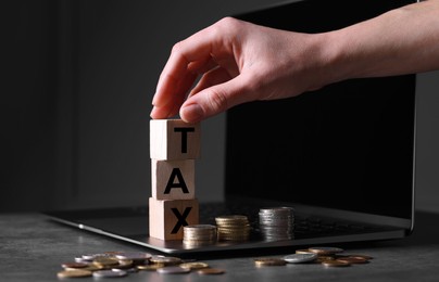Photo of Woman with word Tax made of wooden cubes, laptop and coins at grey table, closeup