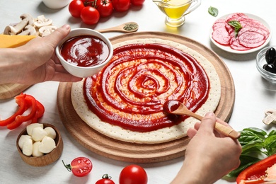 Photo of Woman spreading tomato sauce onto pizza crust and ingredients on white wooden table, closeup