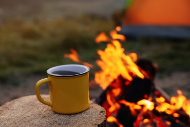 Photo of Yellow mug with hot drink on wooden stump near bonfire outdoors, space for text. Camping season