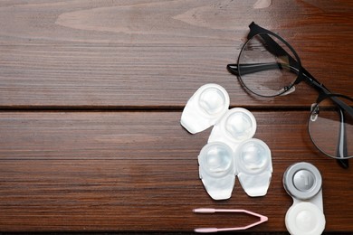 Packages with contact lenses, case, glasses and tweezers on wooden table, flat lay. Space for text
