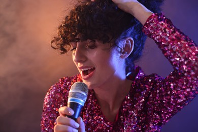 Photo of Beautiful young woman with microphone singing on color background in neon lights and smoke