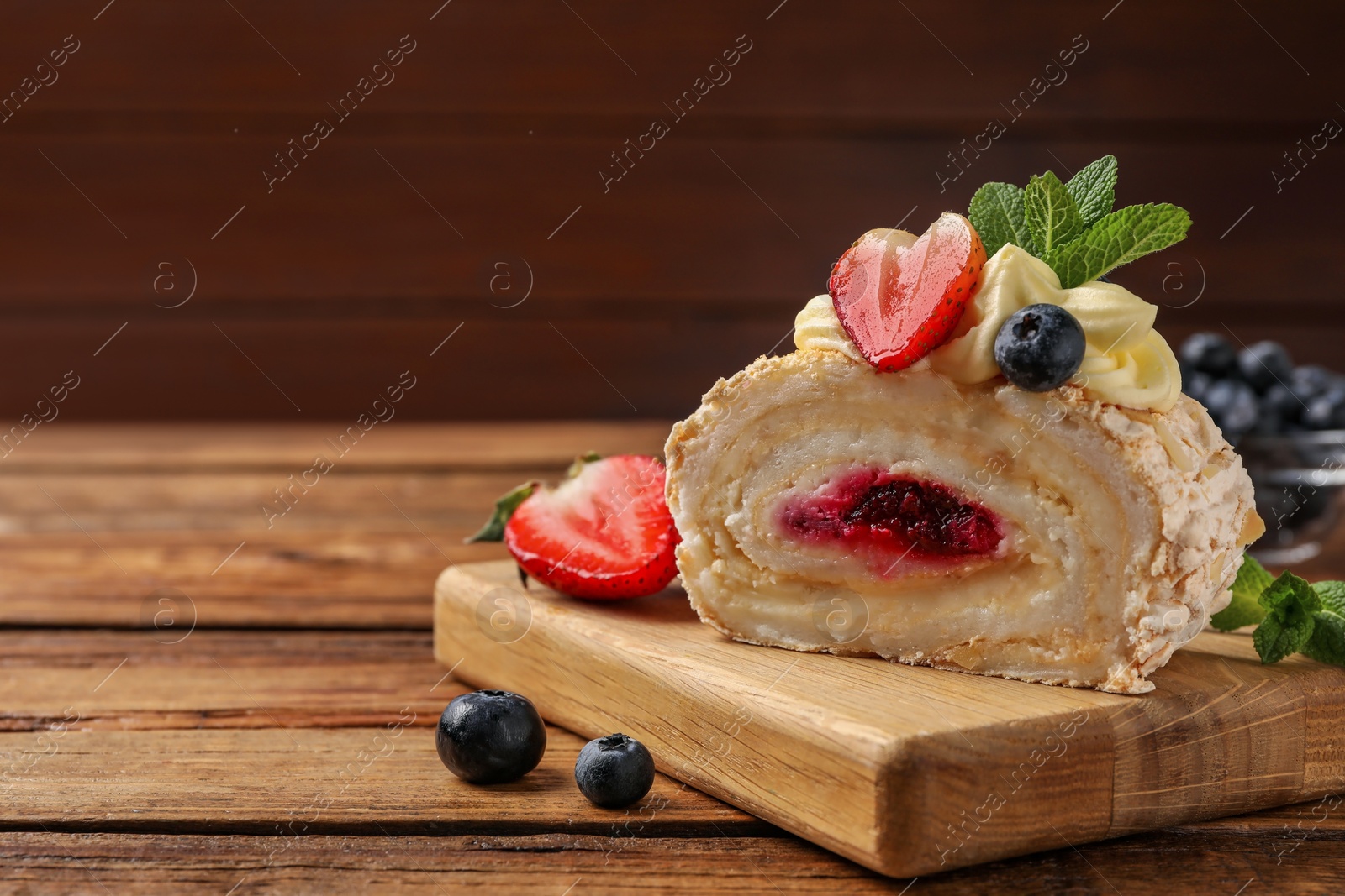 Photo of Piece of tasty meringue roll with jam, cream, strawberry, blueberry and mint on wooden table, closeup. Space for text